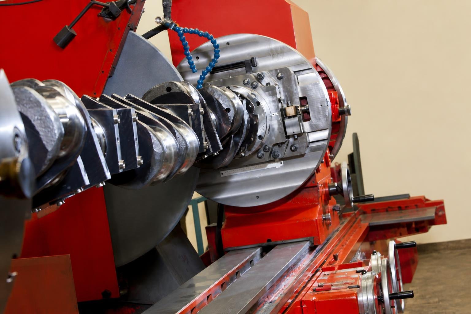 Safety-equipped technician operates a large red crankshaft grinder with a long multi-journal crank mounted horizontally in a heavy engineering workshop.
